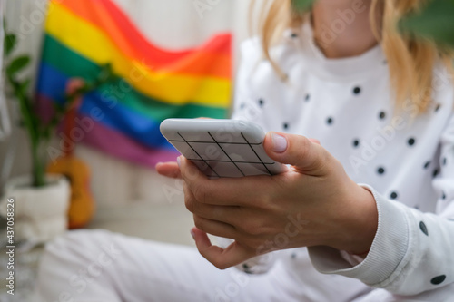 Young millennial hippie woman sitting on balcony using mobile phone. LGBTQ rainbow flag on background. Phone texting, video call, taking selfie. Blogger. Work from home.