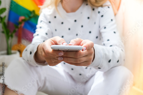 Young millennial hippie woman sitting on balcony using mobile phone. LGBTQ rainbow flag on background. Phone texting, video call, taking selfie. Blogger. Work from home.