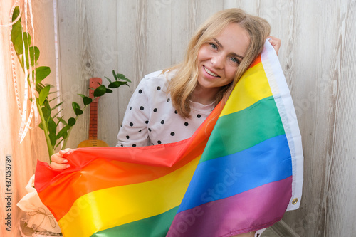 Young blonde millennial hippie woman with rainbow LGBTQ flag at home balcony. Peace and freedom the symbol of LGBT, spending and sharing loving time. Support LGBTQ community.