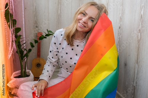 Young blonde millennial hippie woman with rainbow LGBTQ flag at home balcony. Peace and freedom the symbol of LGBT, spending and sharing loving time. Support LGBTQ community.