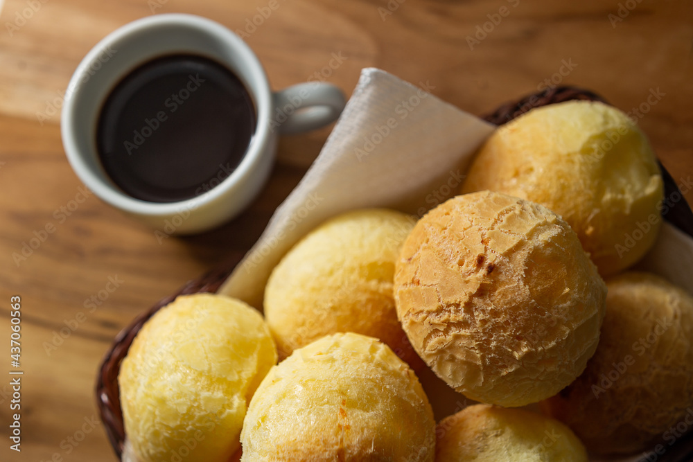 Uma xícara branca cheia de café e uma pequena cesta de vime com pães de queijo sobre uma superfície de madeira. Uma xícara branca cheia de café e uma pequena cesta de vime com pães de queijo sobre uma superfície de madeira.