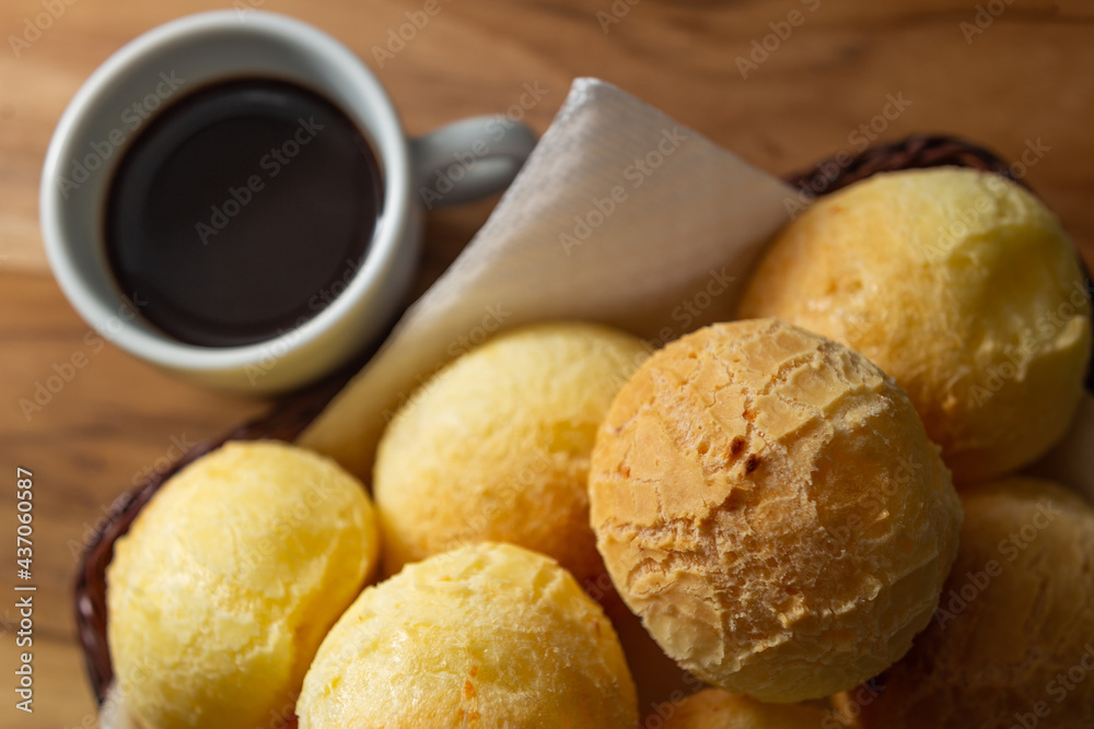 Uma xícara branca cheia de café e uma pequena cesta de vime com pães de queijo sobre uma superfície de madeira. Uma xícara branca cheia de café e uma pequena cesta de vime com pães de queijo sobre uma superfície de madeira.