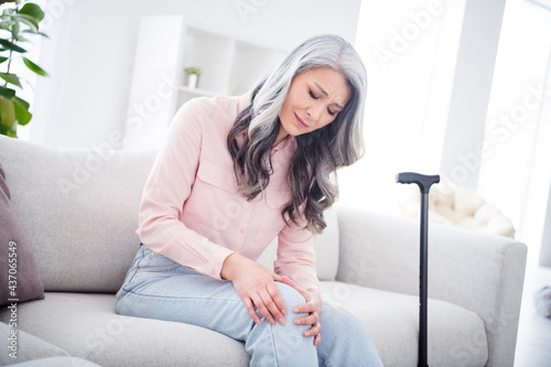 Profile side photo of aged woman unhappy sad upset pain suffer injury knee healthcare meds stick sit couch home photo