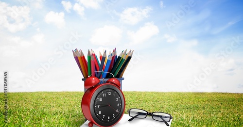 Composition of coloured pencils in pot with glasses and alarm clock, in sunny field with blue sky