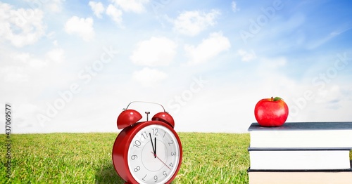 Composition of red retro alarm clock with apple on stack of books over grass