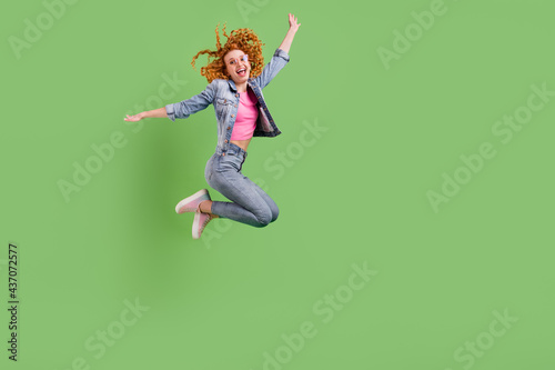 Full length photo of playful sweet young woman wear jeans outfit spectacles jumping empty space smiling isolated green color background