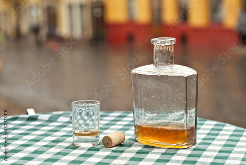 Bottle of alcohol near a glass on an empty table. The concept of alcoholism, loneliness and depression. Old vintage bottle on a checkered tablecloth. Blurred city street on background.