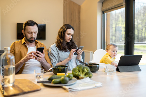 Young parents and their one year baby boy sitting in mobile devices separately during lunch at home. The problem of Internet addiction of parents and raising children