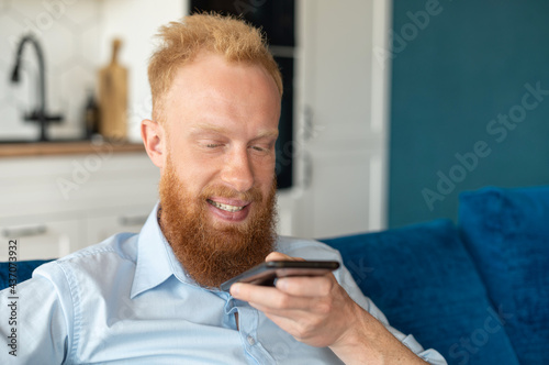 Charismatic redhead guy record voice message and smiling, sits on the sofa at home and holding smartphone near mouth and talking in the mic, chatting, using voice recognizable mobile application
