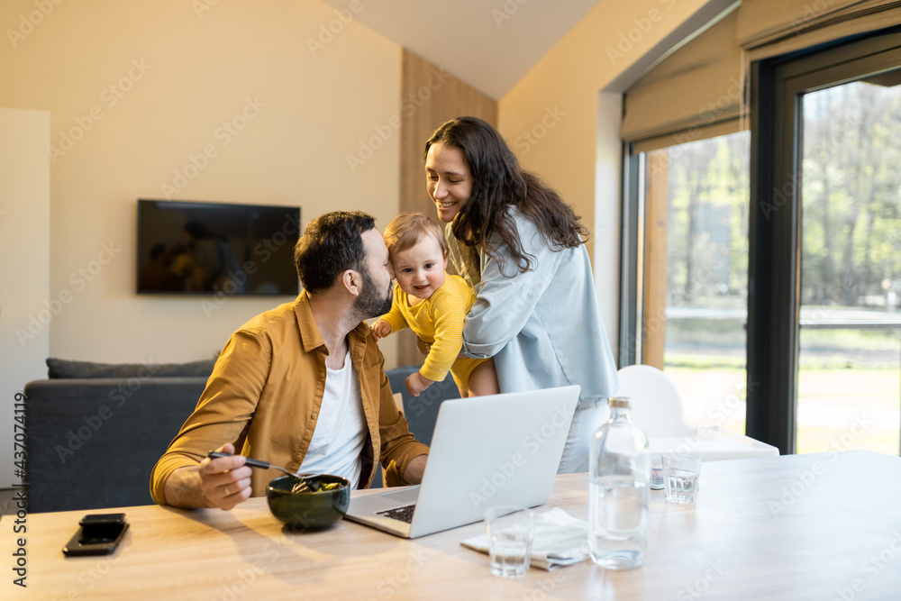 Young father works on a laptop during a lunch time at home with his wife and one year son. Home family routine and work from home concept Young father works on a laptop during a lunch time at home with his wife and one year son. Home family routine and work from home concept