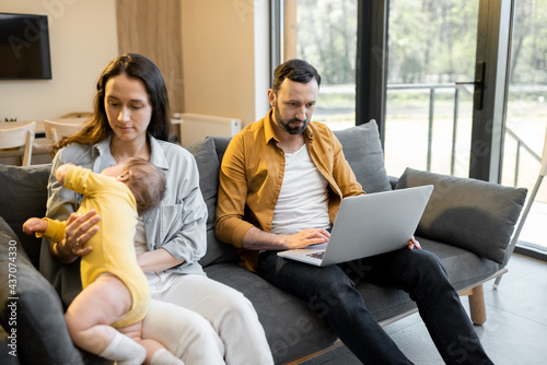 Man works from home with his wife and little son, sitting together on a sofa. Baby interferes father with a work photo