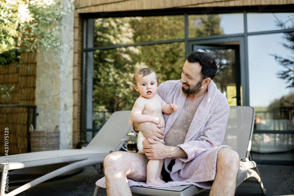 Father in bathrobe with a one year baby boy resting at Spa resort, sitting on a sunbed near the sauna outdoors. Father with a newborn son on vacation Father in bathrobe with a one year baby boy resting at Spa resort, sitting on a sunbed near the sauna outdoors. Father with a newborn son on vacation