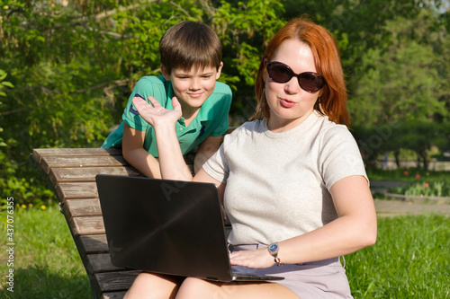 Young red-haired woman in park with her little son. Mom is outraged that her son is interfering with her work. Woman with laptop trying to work in the park and combine work and walk with a child