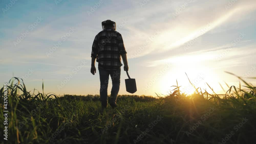 Farm agriculture concept. Farmer with a shovel in rubber boots walks across field at sunset. Silhouette man gardener walking along the road along the field. Tired man after hard work on the ground.