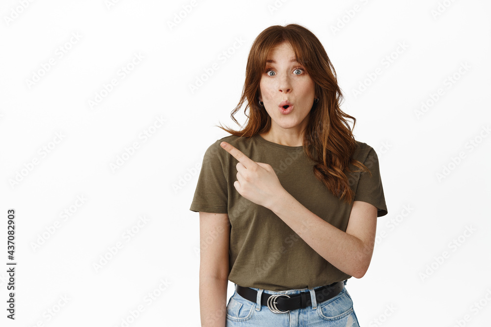 Obraz premium Surprised and excited girl say wow, pointing at upper left corner, showing advertisement with finger, staring in awe at camera, standing against white background