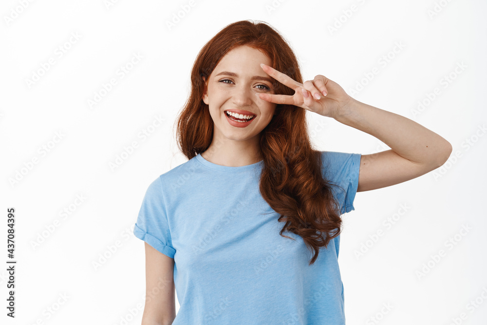 Positive redhead curly girl show peace v-sign and smiling broadly, standing relaxed in casual clothes with cheerful face expression, white background Positive redhead curly girl show peace v-sign and smiling broadly, standing relaxed in casual clothes with cheerful face expression, white background