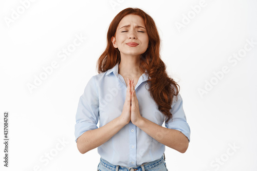 Image of redhead girl begging for help, looking pleading, need favour, supplicating say please, standing in blouse against white background © Cookie Studio