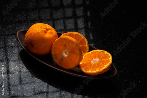 One whole tangerine and one cut in a brown canoe-shaped dish on a black stone table with a window reflection