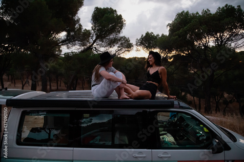 Two women sit down on the roof of a camper van in the forest at sunset © Manu Reyes