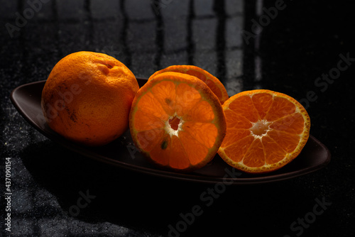 One whole tangerine and one cut in a brown canoe-shaped dish on a black stone table with a window reflection