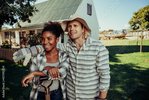 Diverse couple friends smiling into distance showing affection standing in farm village in front of white home dressed in gardening gear  photo