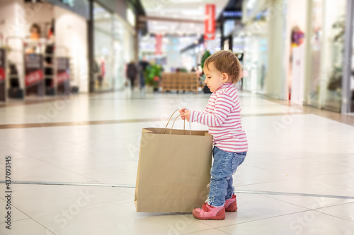 Black friday sale concept, shopping with children, nature friendly shopping. little cute baby girl picks up big craft paper bag for shopping. close-up, soft focus, in the background shops, side view