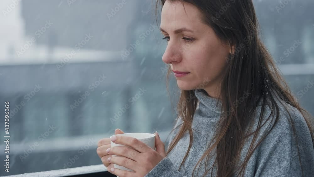 Caucasian woman stays on balcony during snowfall with cup of hot coffee or tea. She looks at the snowflakes and breathes in the frosty fresh air Caucasian woman stays on balcony during snowfall with cup of hot coffee or tea. She looks at the snowflakes and breathes in the frosty fresh air