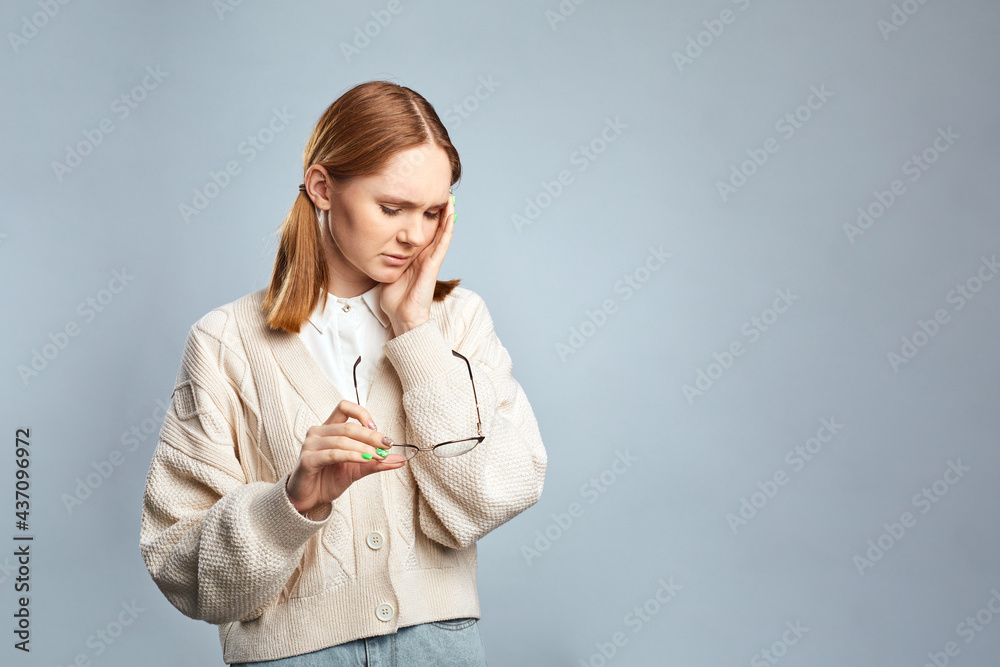 Young tired ill student girl in beige sweater hold glasses put hand on face having headache isolated on gray background. Stressed woman feeling tired after night without sleep. Young tired ill student girl in beige sweater hold glasses put hand on face having headache isolated on gray background. Stressed woman feeling tired after night without sleep.