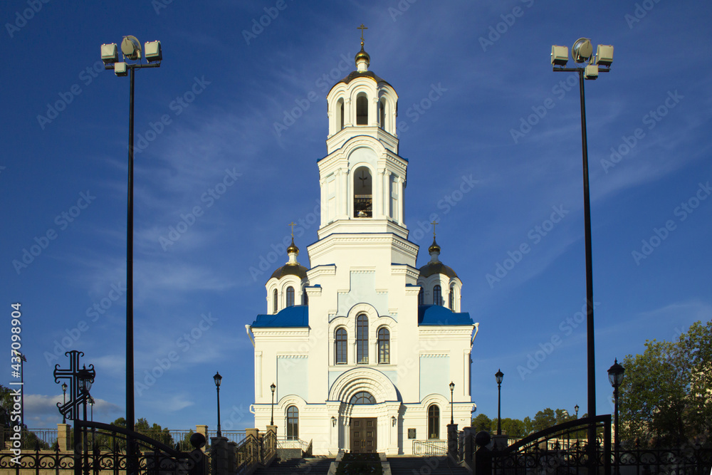 White Orthodox church with three golden domes against a blue sky with stratus clouds. 
The temple is part of the temple complex. A wide staircase leads to it. In the foreground, on both sides