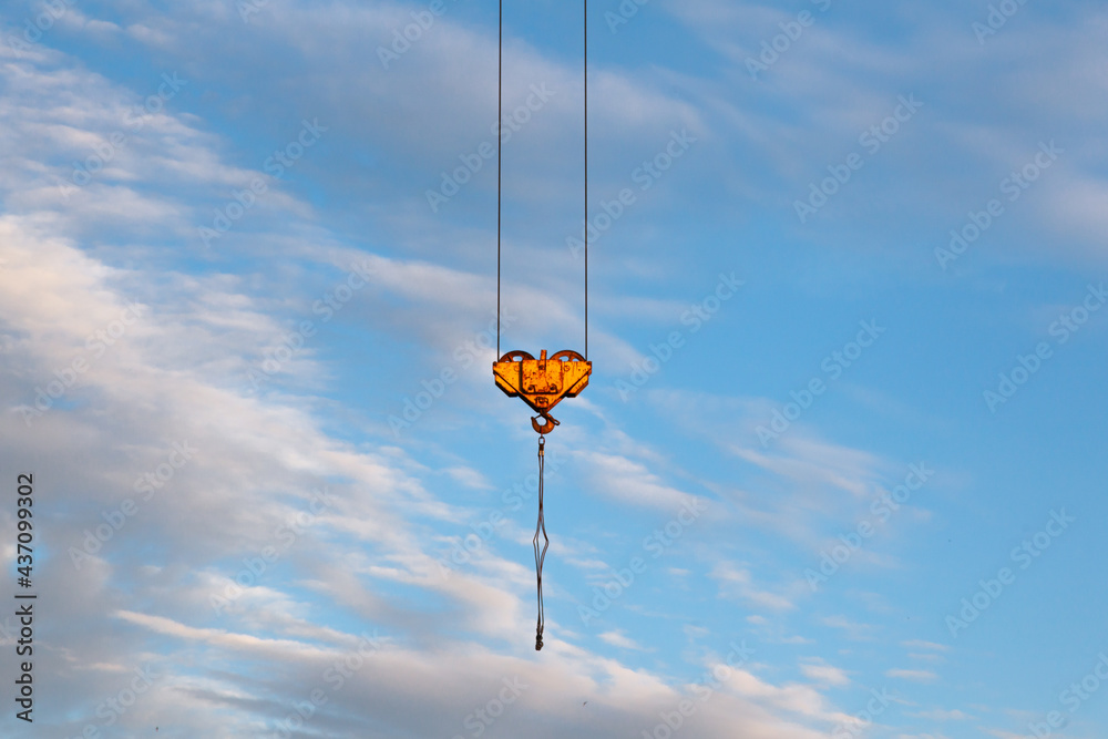 A load hook with rollers of a tower crane with a steel cable at the end against a blue sky with clouds. A load hook with rollers of a tower crane with a steel cable at the end against a blue sky with clouds.