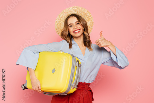 Cheerful woman in sun hat holding suitcase and showing like gesture isolated on pink