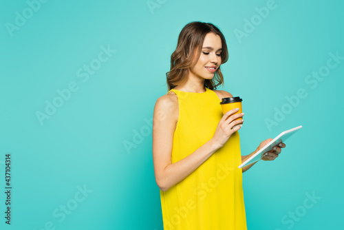 Smiling woman holding paper cup and digital tablet isolated on blue