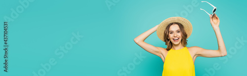 Positive woman with straw hat and sunglasses isolated on blue, banner