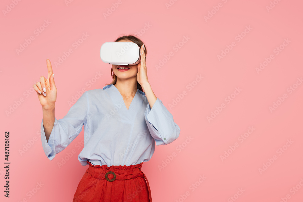 Smiling woman in vr headset pointing with finger isolated on pink Smiling woman in vr headset pointing with finger isolated on pink