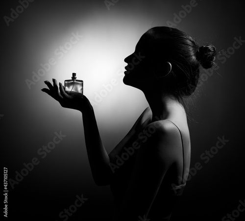 The silhouette of a beautiful young woman in profile with a bottle of perfume in her hand on a dark background.