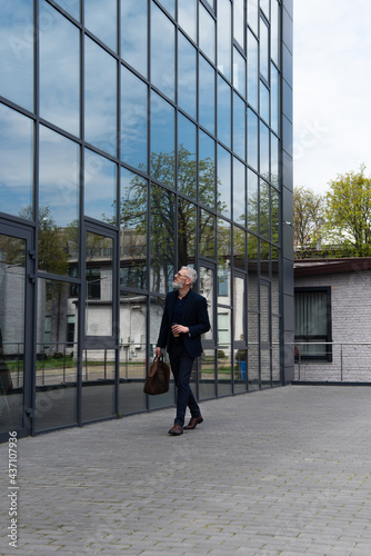 full length of businessman with grey hair holding leather bag and paper cup while walking near modern building © LIGHTFIELD STUDIOS