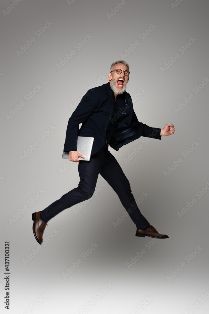 full length of excited man with grey hair holding laptop while levitating on grey