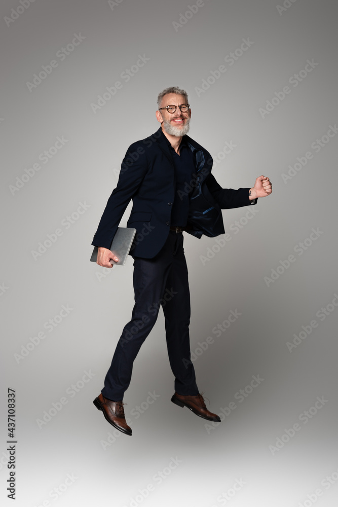 full length of cheerful man with grey hair holding laptop while levitating on grey full length of cheerful man with grey hair holding laptop while levitating on grey