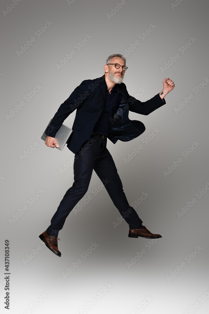 full length of happy man with grey hair holding laptop while levitating on grey