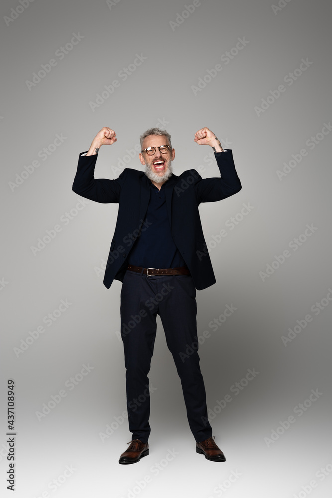 full length of excited middle aged man in glasses and suit rejoicing on grey full length of excited middle aged man in glasses and suit rejoicing on grey