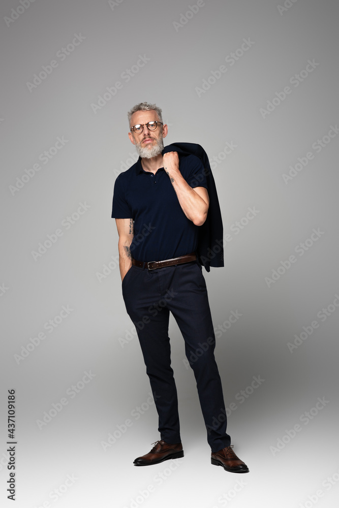 full length of tattooed businessman in glasses and suit posing with hand in pocket on grey full length of tattooed businessman in glasses and suit posing with hand in pocket on grey