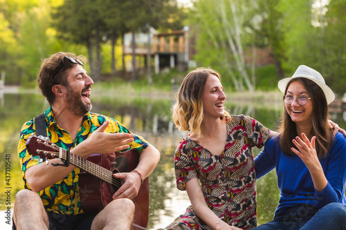 Group of happy friends chatting and laughing together, playing guitar and relaxing. Best friends having fun by the lake. Concept about lifestyle, friendship and people.