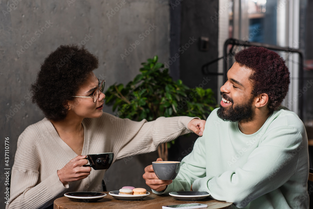 Side view of smiling african american man with coffee sitting near angry girlfriend in cafe