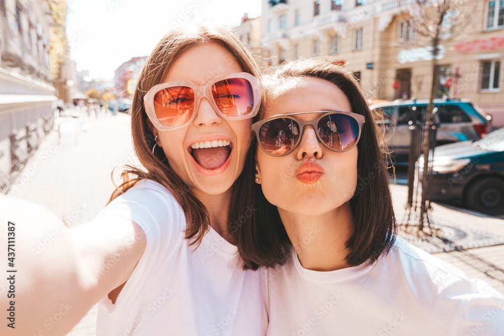 Two young beautiful smiling hipster female in trendy summer white t-shirt clothes and jeans.Sexy carefree women posing on street background.Positive models having fun, hugging and taking Pov selfie
