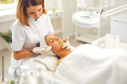 Microdermabrasion skincare in beauty salon. Smiling woman dermatologist making skincare procedure of ultrasound Microdermabrasion with special metal machine for relaxing young woman photo