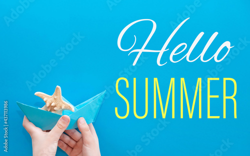 Hello summer. Summer card with a paper boat and a starfish on a blue background in the hands of a child. Travel to the sea and vacation in summer