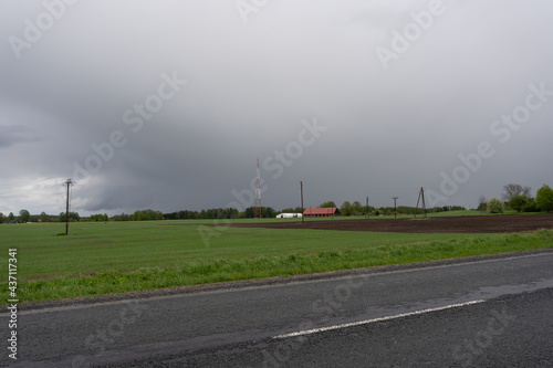 wet asphalt road behind which is a green meadow where in the distance you can see the red roof of the building above which there are dark rain clouds