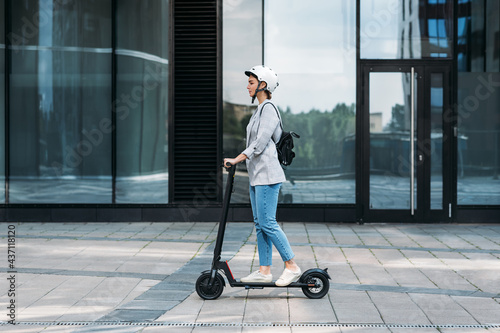 Side view of a young businesswoman riding electric push scooter. Female in cycling helmet driving electrical scooter against building. photo