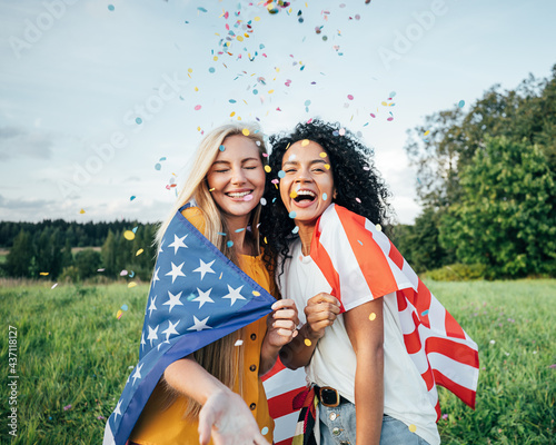Two happy women covered by the USA flag. Young friends celebrating the 4th of July throwing confetti on a field.