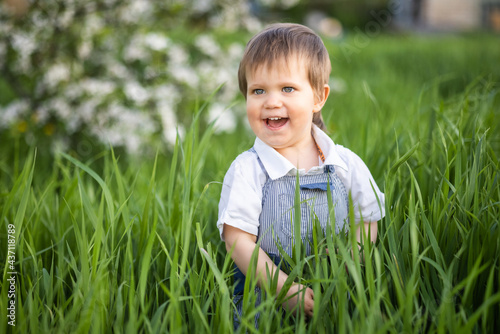 A cute boy with expressive blue eyes in a fashionable jumpsuit smiles funny and hides in the tall green grass